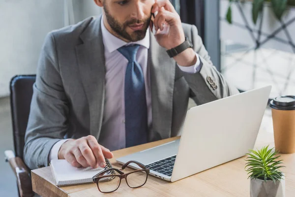 Colpo ritagliato di uomo d'affari che parla da smartphone e utilizzando il computer portatile in ufficio — Foto stock