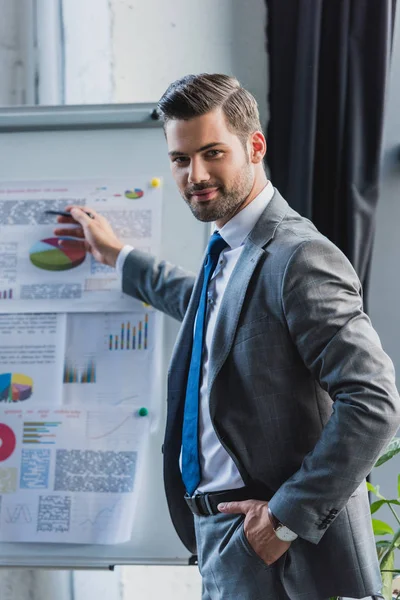 Confident young businessman pointing at whiteboard with business charts and smiling at camera — Stock Photo