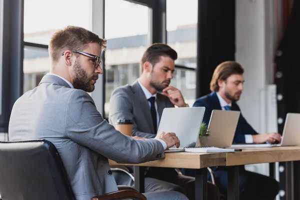 Tres jóvenes empresarios enfocados en el uso de computadoras portátiles en la oficina - foto de stock