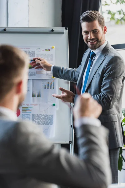 Selective focus of smiling young businessman pointing at whiteboard with business charts and looking at colleague sitting on foreground — Stock Photo