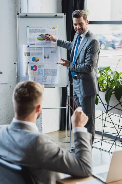 Lächelnder junger Geschäftsmann zeigt auf Whiteboard mit Wirtschaftsdiagrammen und blickt auf Kollegen, die im Vordergrund sitzen — Stockfoto