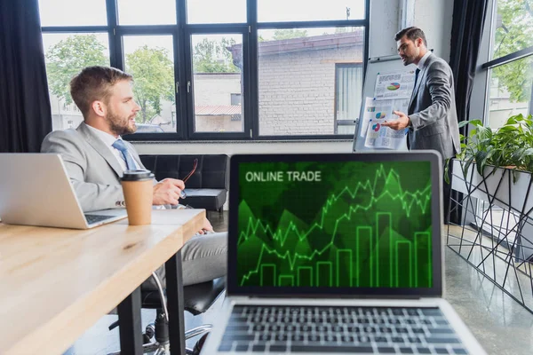 Close-up view of laptop with online trade graphs on screen and businessmen discussing project behind — Stock Photo