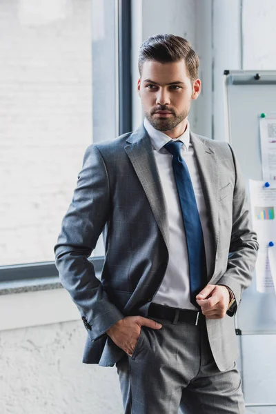 Handsome serious young businessman in suit standing with hand in pocket in office — Stock Photo