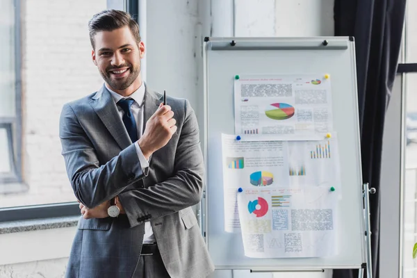 Handsome young businessman smiling at camera while standing near whiteboard with business charts in office — Stock Photo