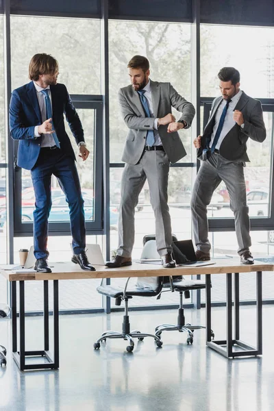 Tres jóvenes empresarios sonrientes bailando en la mesa en la oficina - foto de stock