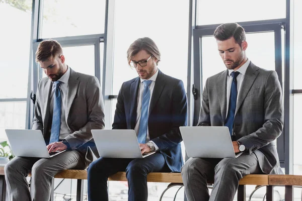 Tres jóvenes hombres de negocios en trajes sentados y usando computadoras portátiles - foto de stock