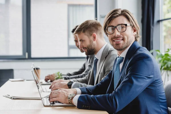 Joven hombre de negocios en gafas de vista utilizando el ordenador portátil y sonriendo a la cámara mientras trabaja con colegas en la oficina - foto de stock