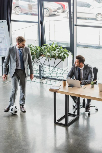 Vue grand angle de jeune homme d'affaires jouant avec le ballon de football et regardant un collègue souriant en utilisant un ordinateur portable au bureau — Photo de stock