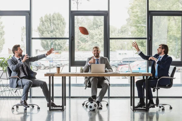 Smiling young businessmen using laptops and playing with soccer and rugby balls in office — Stock Photo