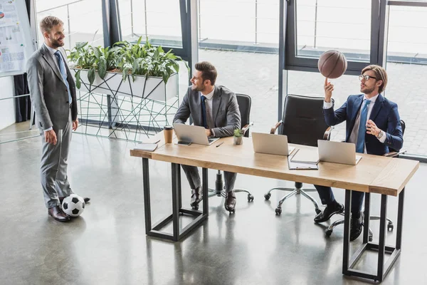 Vue à grand angle de jeunes hommes d'affaires utilisant des ordinateurs portables et jouant avec des ballons de football et de basket au bureau — Photo de stock