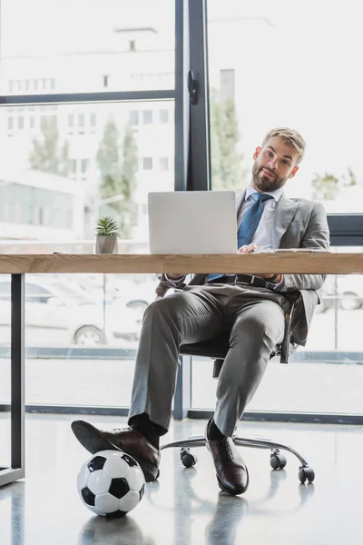 Jeune homme d'affaires avec ballon de football en utilisant un ordinateur portable et souriant à la caméra dans le bureau — Photo de stock