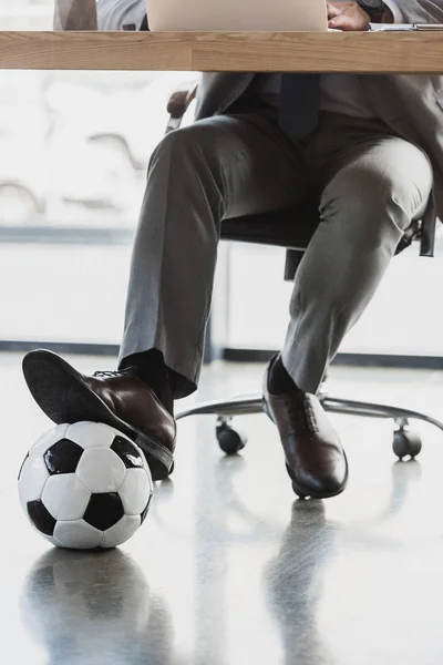 Recortado tiro de joven hombre de negocios con pelota de fútbol usando portátil en la oficina - foto de stock