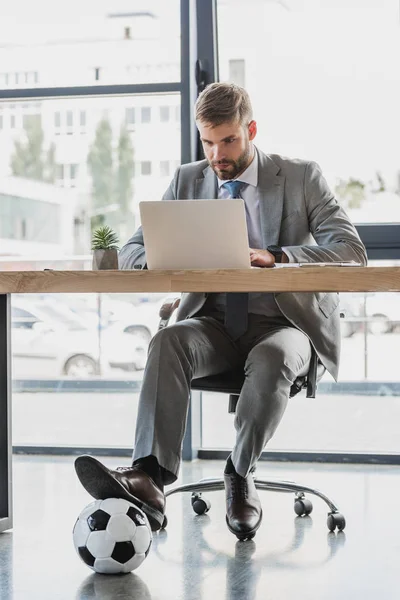 Jeune homme d'affaires avec ballon de football en utilisant un ordinateur portable au bureau — Photo de stock