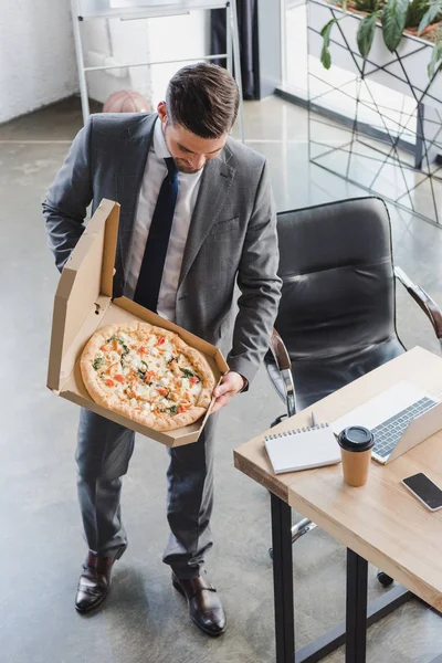 Vue grand angle de beau jeune homme d'affaires en costume tenant boîte avec pizza au bureau — Photo de stock