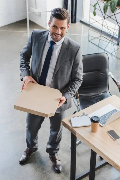 Visão de alto ângulo de belo jovem empresário em terno segurando caixa de pizza e sorrindo para a câmera no escritório — Fotografia de Stock