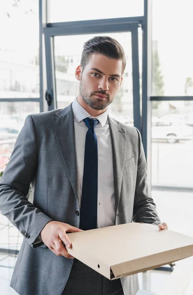 Красивий молодий бізнесмен у костюмі тримає коробку для піци і дивиться на камеру в офісі — стокове фото
