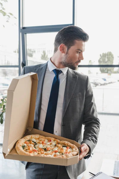 Bonito jovem empresário no terno segurando caixa com pizza no escritório — Fotografia de Stock