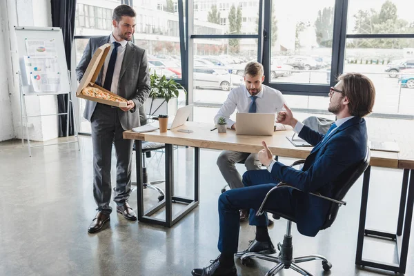 Smiling young businessman holding pizza in box and looking at coworkers in office — Stock Photo