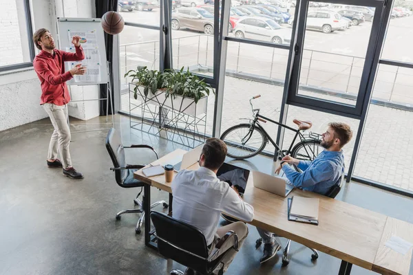 Високий кут зору молодих бізнесменів, які грають з баскетбольним м'ячем і використовують ноутбуки в офісі — стокове фото