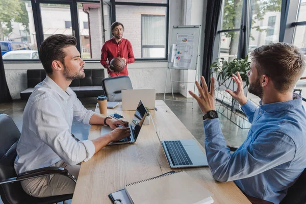 Jeunes hommes d'affaires jouant avec le ballon de basket tout en utilisant des ordinateurs portables au bureau — Photo de stock