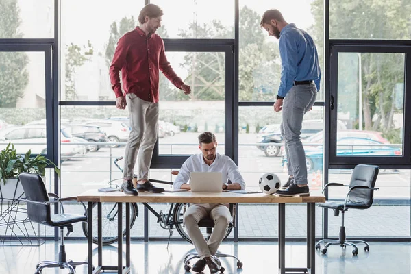 Jovem empresário usando laptop enquanto colegas jogando com bola de futebol na mesa — Fotografia de Stock