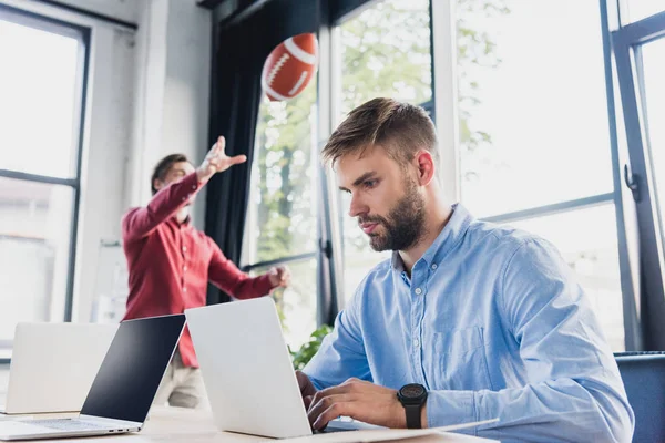 Focado jovem empresário usando laptop enquanto colega jogando com bola de rugby atrás no escritório — Fotografia de Stock