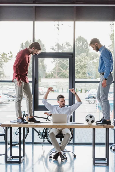 Triunfante hombre de negocios utilizando el ordenador portátil, mientras que los colegas jugando con la pelota de fútbol en la mesa - foto de stock