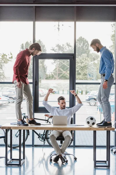 Triumphing businessman using laptop while colleagues playing with soccer ball on table — Stock Photo