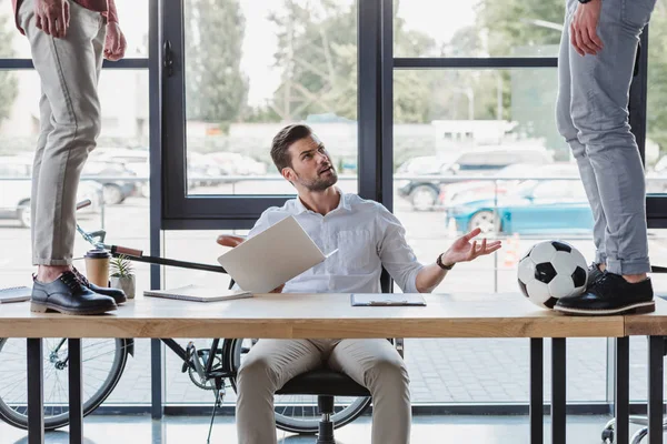 Partial view of men kicking soccer ball on table while irritated colleague using laptop in office — Stock Photo