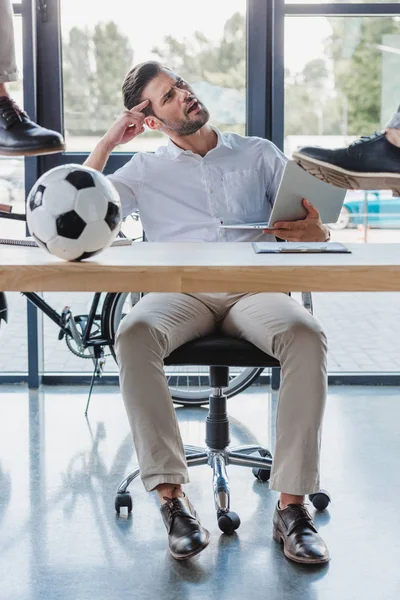 Cropped shot of men kicking soccer ball on table while angry colleague using laptop in office — Stock Photo