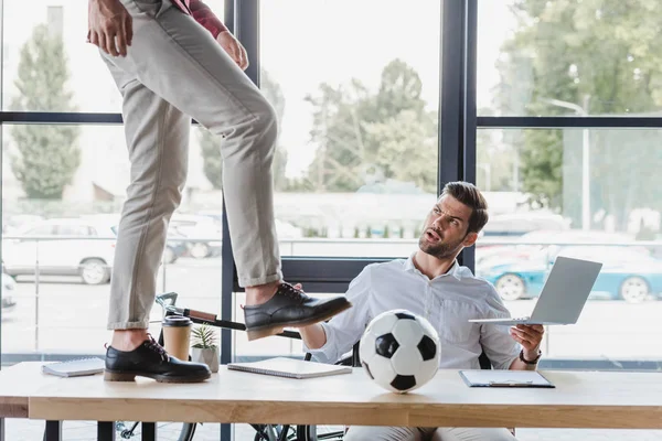 Recortado tiro de hombre pateando pelota de fútbol en la mesa mientras colega usando portátil en la oficina - foto de stock