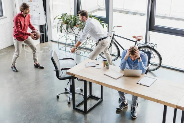 Vue à angle élevé de jeunes hommes d'affaires s'amusant avec le ballon de basket-ball tout en collègue essayant de travailler au bureau — Photo de stock