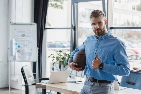 Handsome young businessman holding basketball ball and showing thumb up in office — Stock Photo