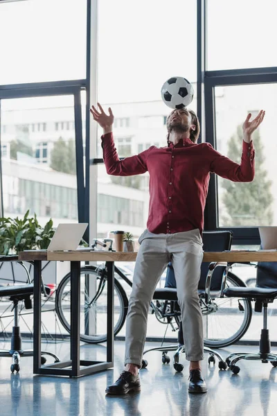 Vista de ángulo bajo del joven empresario que equilibra la pelota de fútbol en la cabeza en la oficina - foto de stock