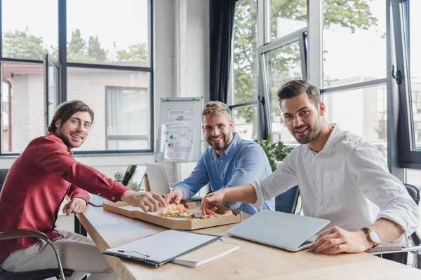 Giovani uomini d'affari che mangiano pizza e sorridono alla telecamera in ufficio — Foto stock
