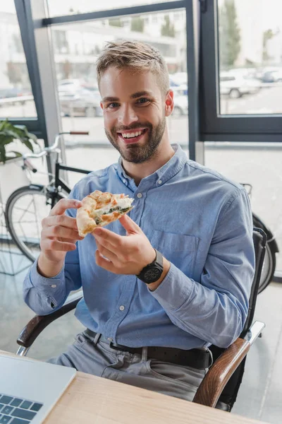 Guapo joven empresario sosteniendo pizza y sonriendo a la cámara en la oficina - foto de stock