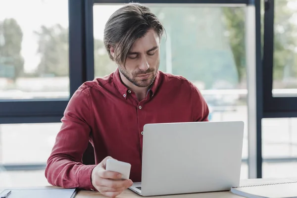 Empresário segurando smartphone e usando laptop enquanto trabalhava no escritório — Fotografia de Stock