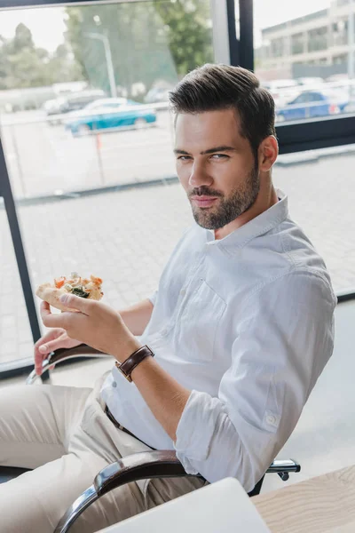 Vue grand angle de jeune homme d'affaires mangeant de la pizza et regardant la caméra — Photo de stock
