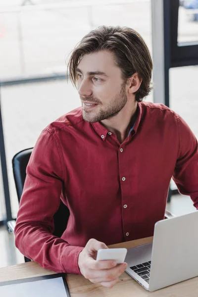Высокий угол зрения красивый улыбающийся молодой бизнесмен смотрит в сторону во время использования смартфона и ноутбука в офисе — стоковое фото