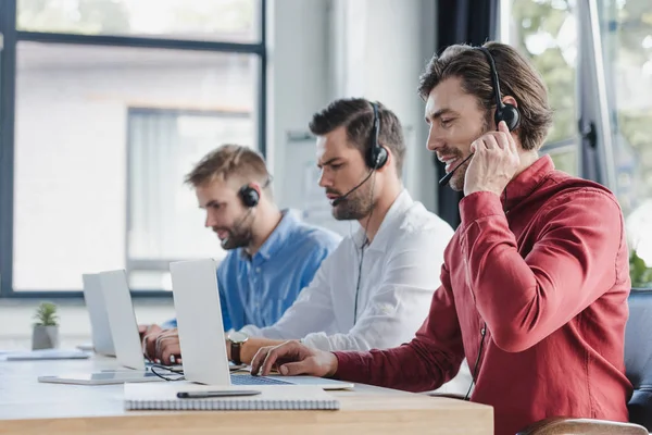 Tres jóvenes operadores de centros de llamadas en auriculares con ordenadores portátiles en la oficina - foto de stock