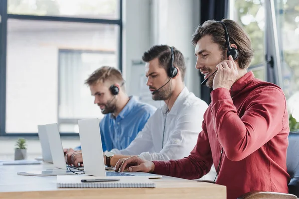 Trois jeunes opérateurs de centre d'appels dans les casques utilisant des ordinateurs portables dans le bureau — Photo de stock