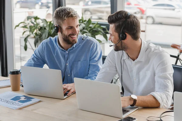 Handsome young call center operators in headsets smiling each other while using laptops in office — Stock Photo