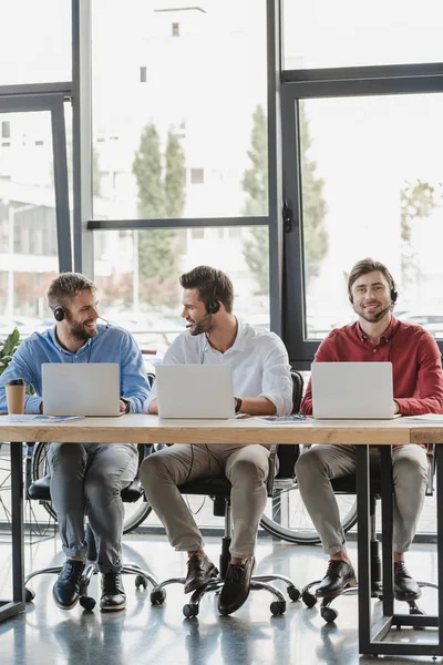 Three smiling young call center operators in headsets using laptops in office — Stock Photo