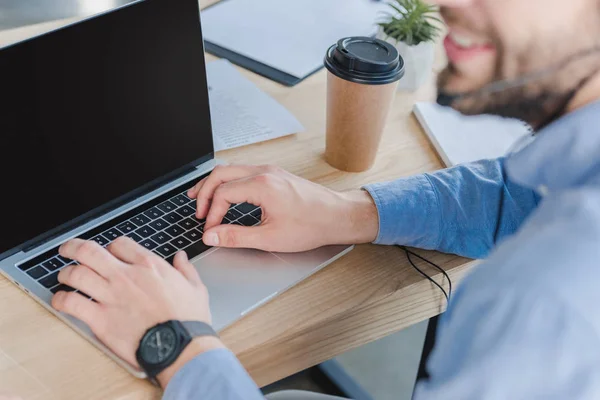 Recortado disparo de sonriente hombre de negocios en auriculares con ordenador portátil con pantalla en blanco en el lugar de trabajo - foto de stock