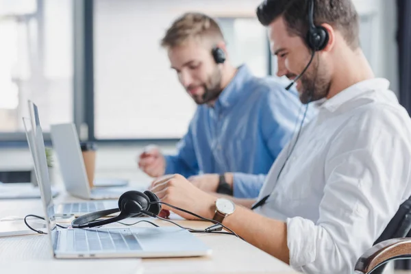 Smiling young call center operators in headsets using laptops in office — Stock Photo