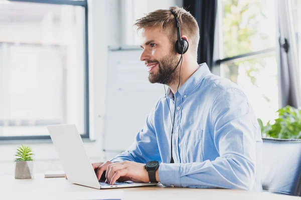 Vista lateral del operador de centro de llamadas joven sonriente en auriculares usando computadora portátil en la oficina - foto de stock