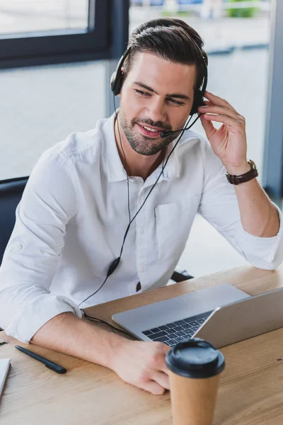 Sonriente operador de centro de llamadas joven en auriculares usando el ordenador portátil en la oficina - foto de stock