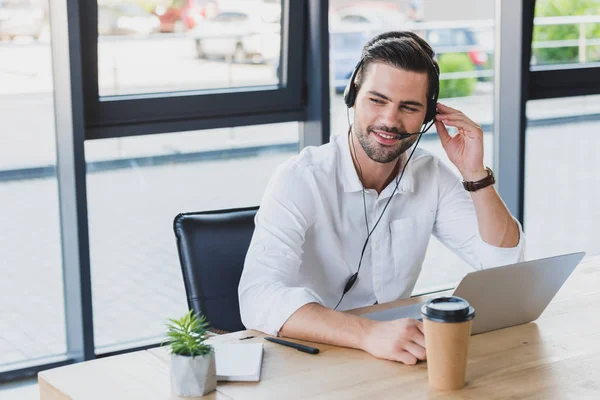 Bonito sorrindo jovem operador de call center no fone de ouvido usando laptop no escritório — Fotografia de Stock