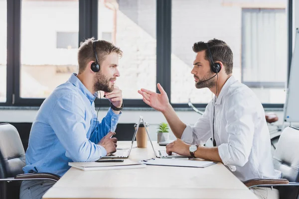 Side view of young call-center operators in headsets discussing and looking at each other while using laptops in office — стоковое фото