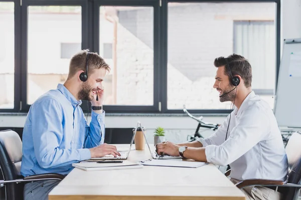 Side view of smiling young call center operators in headsets using laptops in office — Stock Photo